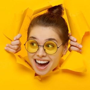 Horizontal banner of young girl in glasses tearing paper and peeking out hole, curious about commercial offer on copy space on left, isolated on yellow background
